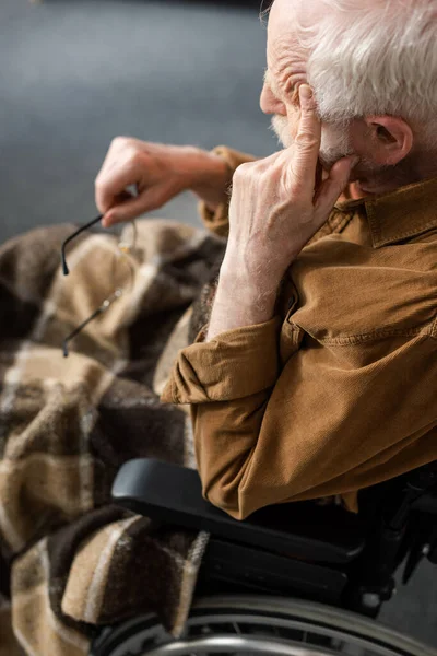 Hombre mayor discapacitado tocando la cara y sosteniendo las gafas mientras está sentado en silla de ruedas - foto de stock