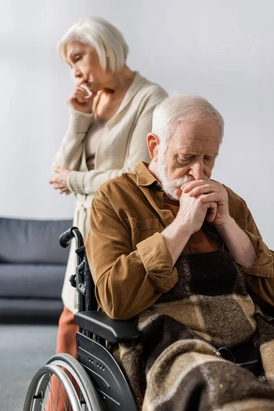 Избирательный фокус грустной старшей жены, стоящей позади инвалида, больного человека, сидящего в инвалидной коляске со сложенными руками — стоковое фото
