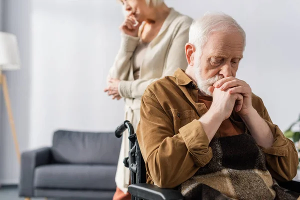 Vista ritagliata di triste donna anziana in piedi vicino a disabili, marito malato seduto in sedia a rotelle con le mani serrate — Foto stock