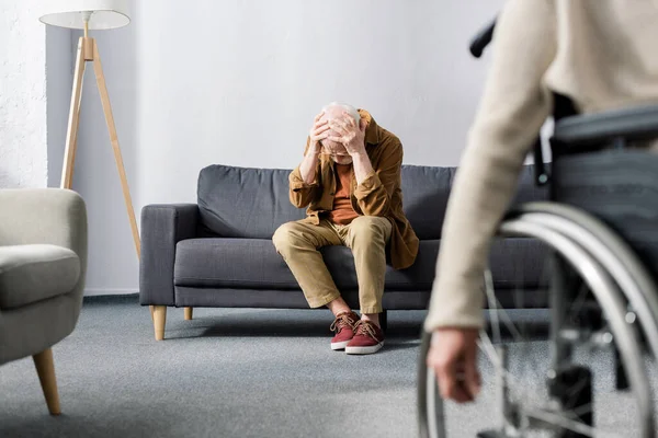 Selektiver Fokus einer behinderten Frau im Rollstuhl und eines älteren, verzweifelten Mannes, der auf dem Sofa sitzt und den Kopf mit den Händen bedeckt — Stockfoto