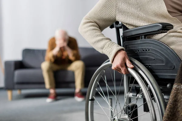 Обрезанный вид женщины-инвалида в инвалидной коляске, и пожилой, отчаявшийся мужчина, сидящий на диване и затемняющий лицо руками — стоковое фото