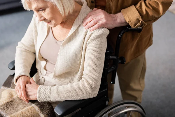 Cropped view of senior man touching shoulder of disabled wife sitting in wheelchair with bowed head — Stock Photo