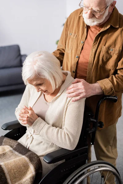 Senior man touching shoulder of disabled wife, sick on dementia, sitting in wheelchair with bowed head and clenched hands — Stock Photo