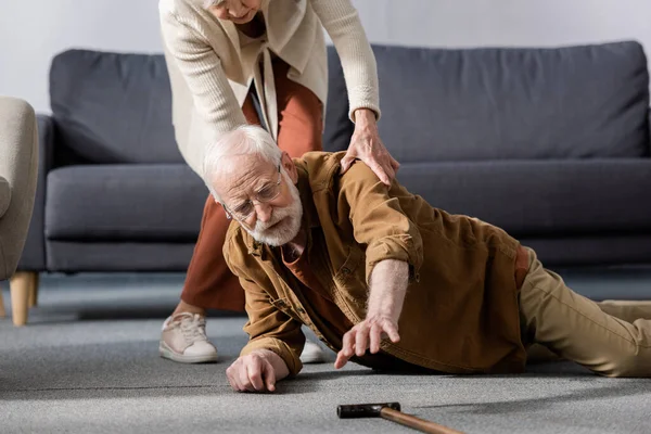 Старший мужчина лежит на полу и пытается получить трость, пока жена помогает ему — стоковое фото