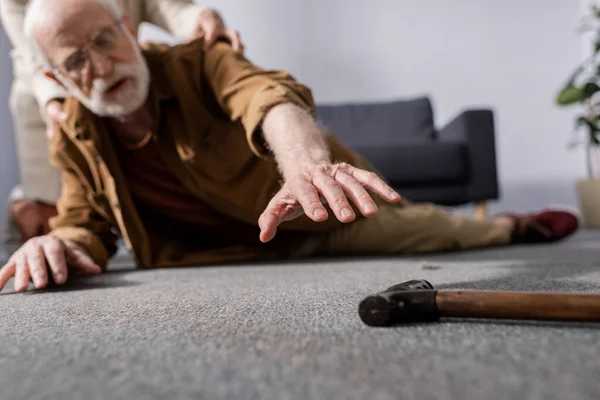 Selective focus of fallen senior man trying to get walking stick while wife helping him — Stock Photo