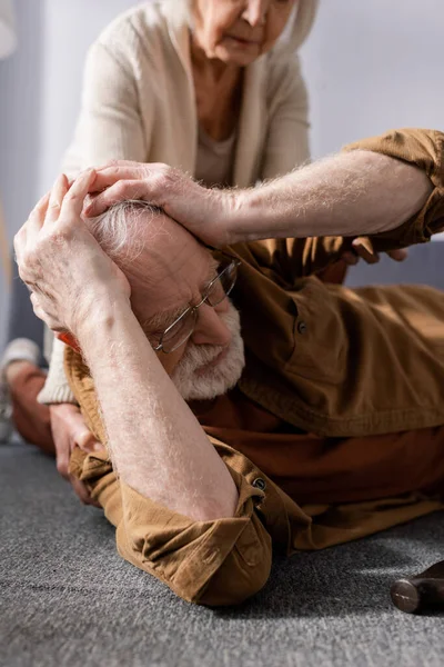 Обрізаний вид на старшу жінку біля чоловіка лежить на підлозі і торкається голови — стокове фото