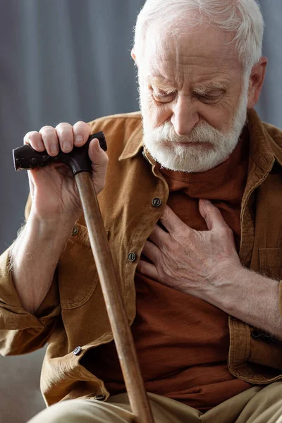 Lonely senior man sitting with walking stick and touching chest while feeling bad — Stock Photo