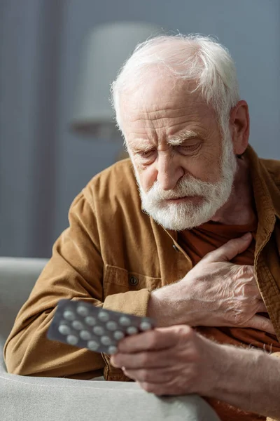 Anciano solitario tocando el pecho mientras se siente mal y sosteniendo pastillas - foto de stock