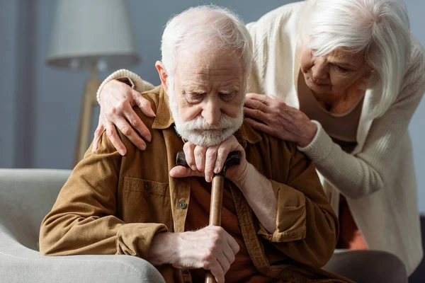 Älterer Mann, an Demenz erkrankt, sitzt mit Gehstock, während seine Frau ihn ansieht und seine Schultern berührt — Stockfoto