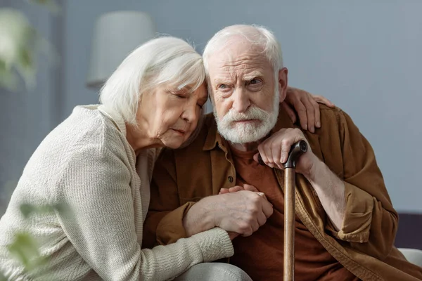Mujer mayor con los ojos cerrados abrazando marido, enfermo de demencia, y sosteniendo su mano - foto de stock