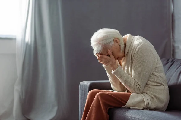 Пригнічена старша жінка сидить на дивані з вклоненим обличчям і затемнює обличчя руками — стокове фото