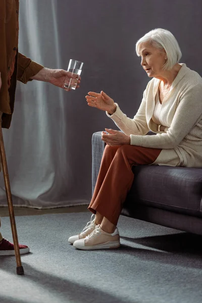 Vue partielle de l'homme âgé donnant un verre d'eau à la femme se sentant mal et assis sur le canapé — Photo de stock