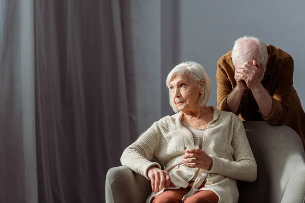 Despaired senior man obscuring face with hand while standing behind sick wife holding glass of water — Stock Photo