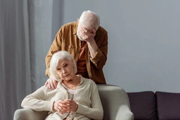 Homme âgé déprimé obscurcissant le visage avec la main tout en se tenant derrière la femme malade tenant un verre d'eau — Photo de stock