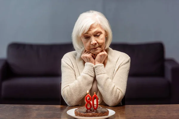 Thoughtful, sad senior woman sitting near birthday cake with number eighty and burning candles alone — Stock Photo
