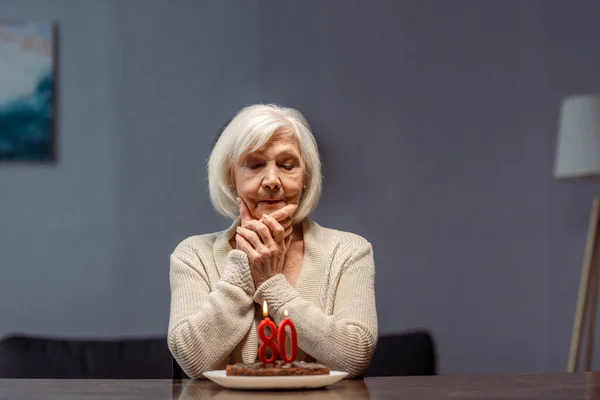 Sênior, mulher solitária tocando rosto enquanto olha para bolo de aniversário com número oitenta e velas acesas — Fotografia de Stock