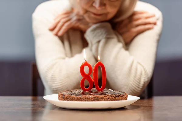 Vista recortada de la mujer mayor sentada sola cerca de bithday pastel con número ochenta y velas encendidas - foto de stock