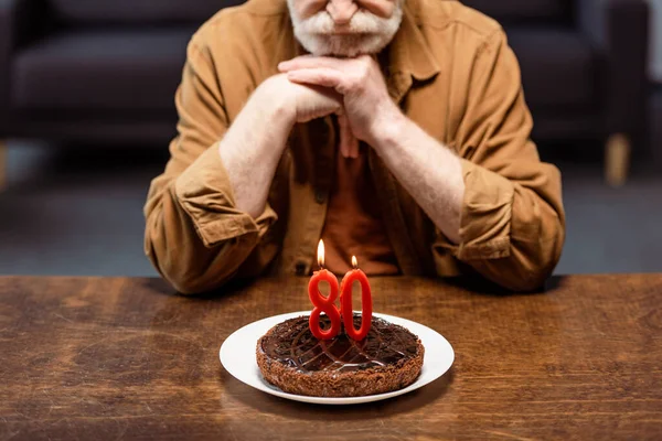 Vista ritagliata di uomo anziano solitario appoggiato su mani serrate e guardando torta di compleanno con il numero ottanta — Foto stock