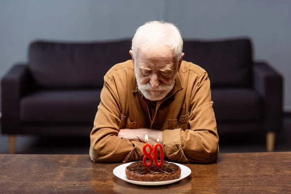 Senior lonely man sitting with crossed arms near birthday cake with number eighty — Stock Photo