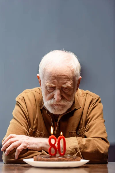 Anziano solitario seduto con le braccia incrociate vicino torta di compleanno con il numero ottanta — Foto stock