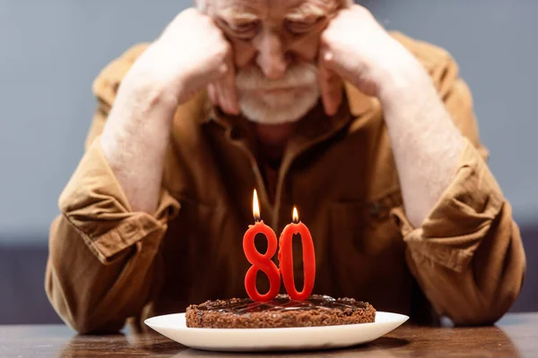 Selective focus of lonely senior man leaning on hands while looking at birthday cake with number eighty — Stock Photo