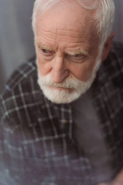 Вибірковий фокус пригніченого старшого чоловіка, який дивиться далеко через віконне скло — стокове фото