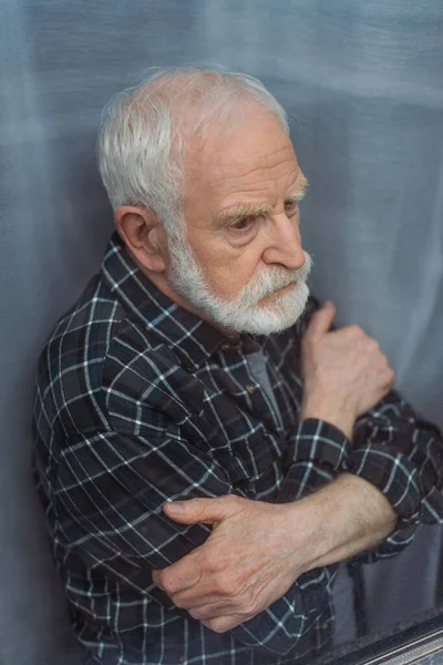 Depressed senior man looking away while standing with crossed arms near window glass — Stock Photo