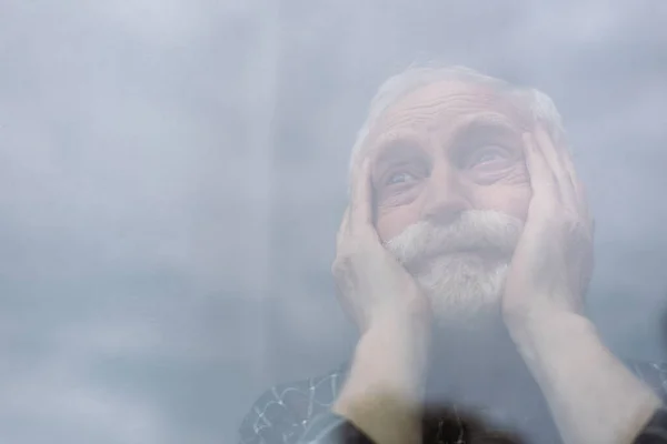 Selective focus of senior, lonely man touching face and looking away through window glass — Stock Photo
