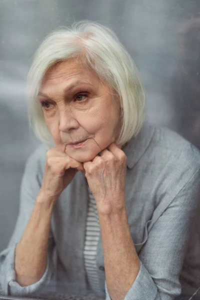 Sad senior woman looking away through window glass — Stock Photo