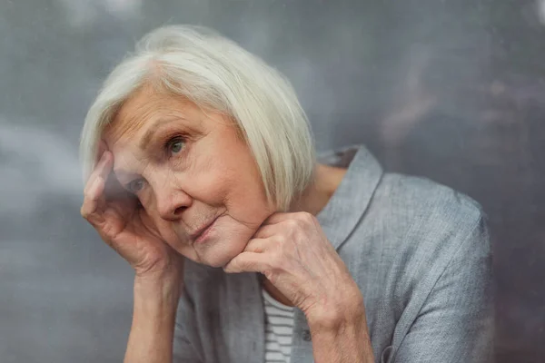 Lonely senior woman loooking away while standing by window — Stock Photo
