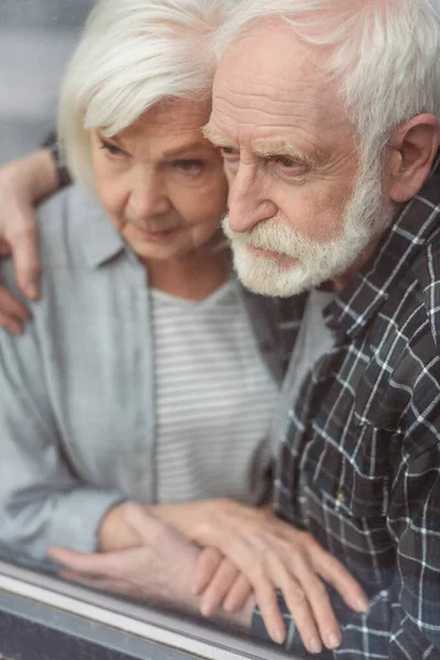 Nachdenklicher Senior und seine an Demenz erkrankte Frau halten Händchen, während sie am Fenster stehen — Stockfoto