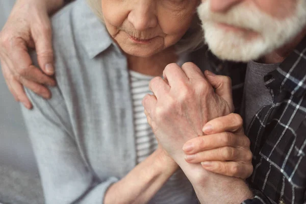 Vista parcial del hombre mayor y su esposa, enfermos de demencia, tomados de la mano mientras están de pie junto a la ventana — Stock Photo