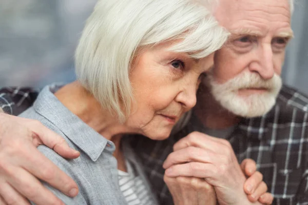 Nachdenklicher älterer Mann hält an der Hand seiner demenzkranken Frau, während er durch das Fenster schaut — Stockfoto