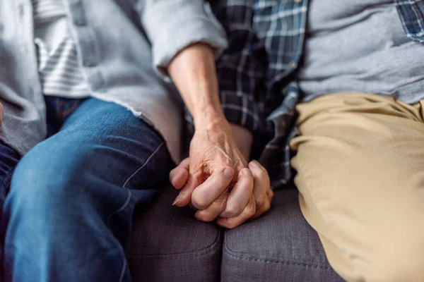 Cropped view of senior couple sitting on sofa and holding hands — Stock Photo