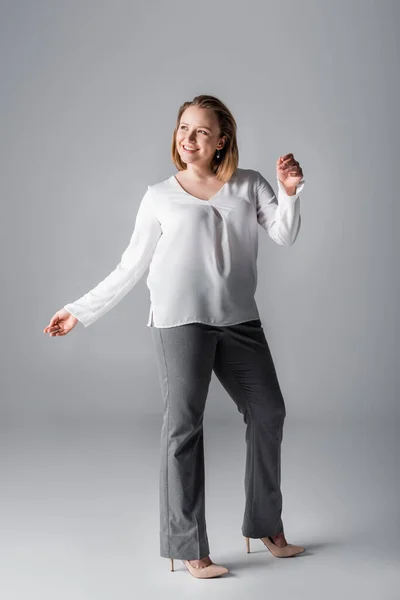 Ansicht des stilvollen übergewichtigen Mädchens, das wegschaut und lächelt, während es auf grau posiert — Stockfoto