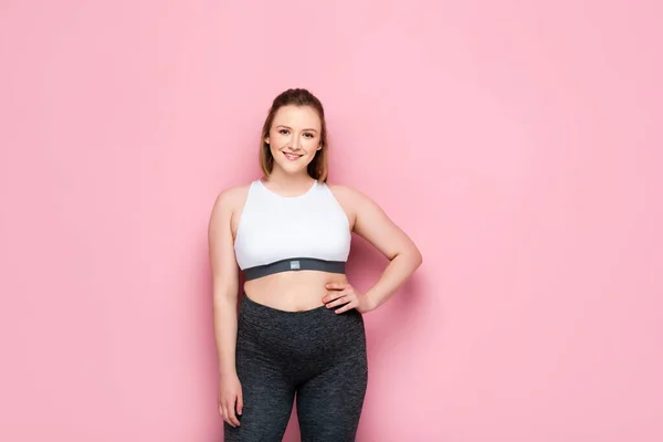 Bella ragazza in sovrappeso in abbigliamento sportivo tenendosi per mano sul fianco e sorridendo sul rosa — Foto stock