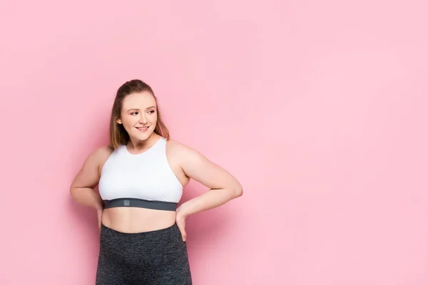 Joyeuse fille en surpoids en vêtements de sport tenant la main sur les hanches et regardant loin sur rose — Photo de stock