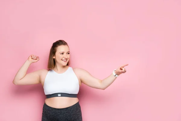 Cheerful overweight girl pointing with finger and looking away on pink — Stock Photo