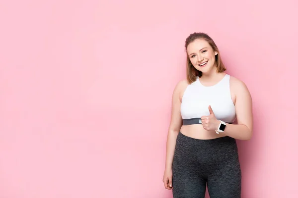 Feliz chica con sobrepeso mostrando el pulgar hacia arriba mientras sonríe a la cámara en rosa - foto de stock