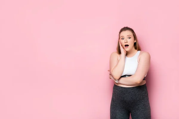 Schockiertes übergewichtiges Mädchen berührt Kinn, während es in die Kamera auf rosa schaut — Stockfoto