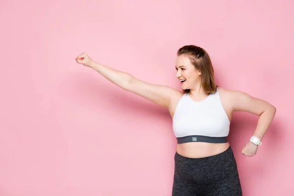 Excitada chica con sobrepeso mostrando el gesto ganador mientras mira hacia otro lado en rosa - foto de stock