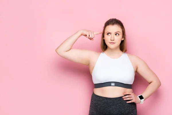 Thoughtful overweight girl pointing with finger at her head on pink — Stock Photo