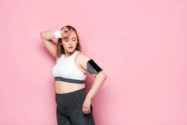 Chica cansada con sobrepeso mirando el teléfono inteligente en el brazalete mientras toca la frente en rosa — Stock Photo