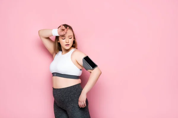 Tired overweight girl looking at smartphone in armband while touching forehead on pink — Stock Photo