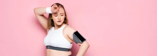 Горизонтальне зображення втомленої дівчини з надмірною вагою, яка дивиться на смартфон у пов'язці, торкаючись лоба на рожевому — стокове фото