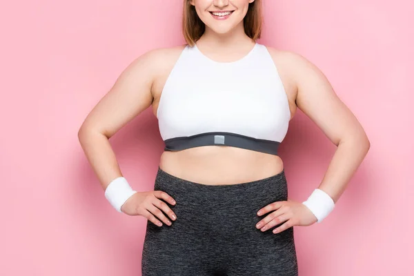 Abgeschnittene Ansicht eines lächelnden übergewichtigen Mädchens in Sportbekleidung, die Hände auf Hüften auf rosa — Stockfoto