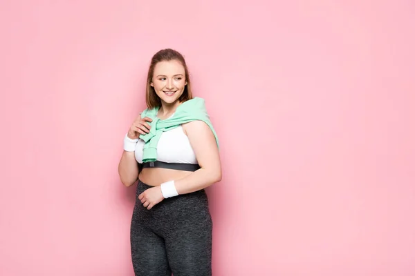 Cheerful overweight girl with sweatshirt over shoulders looking away on pink — Stock Photo
