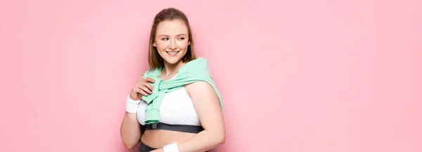 Cultivo panorámico de hermosa chica con sobrepeso con sudadera sobre los hombros sonriendo mientras mira hacia otro lado en rosa - foto de stock