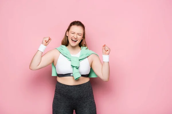 Menina sobrepeso animado com camisola sobre ombros mostrando gesto vencedor com olhos fechados em rosa — Fotografia de Stock