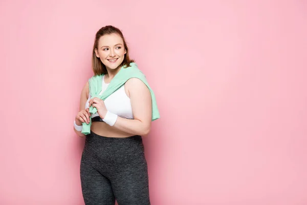 Pretty overweight girl with sweatshirt over shoulders smiling while looking away on pink — Stock Photo